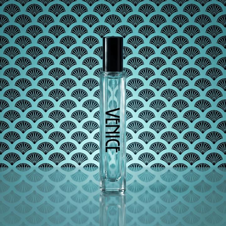 A clear cylindrical perfume bottle with a black cap stands centered against a turquoise background with a repeating black fan pattern. The bottle has the brand name 'VENICE' printed vertically in black.
