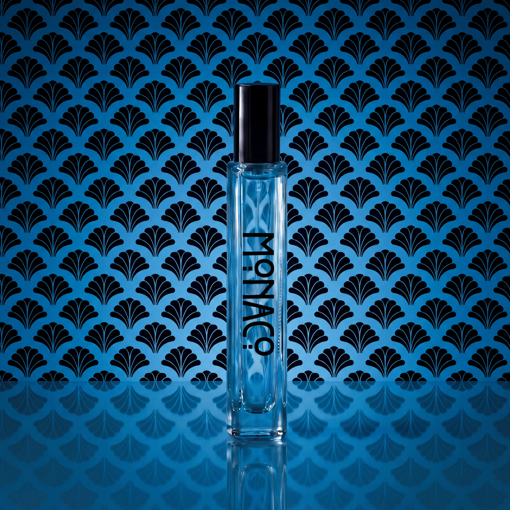 A clear rectangular perfume bottle with a black cap stands centered against a blue background with a repeating black fan-like pattern. The bottle is labeled 'MONACO' vertically.