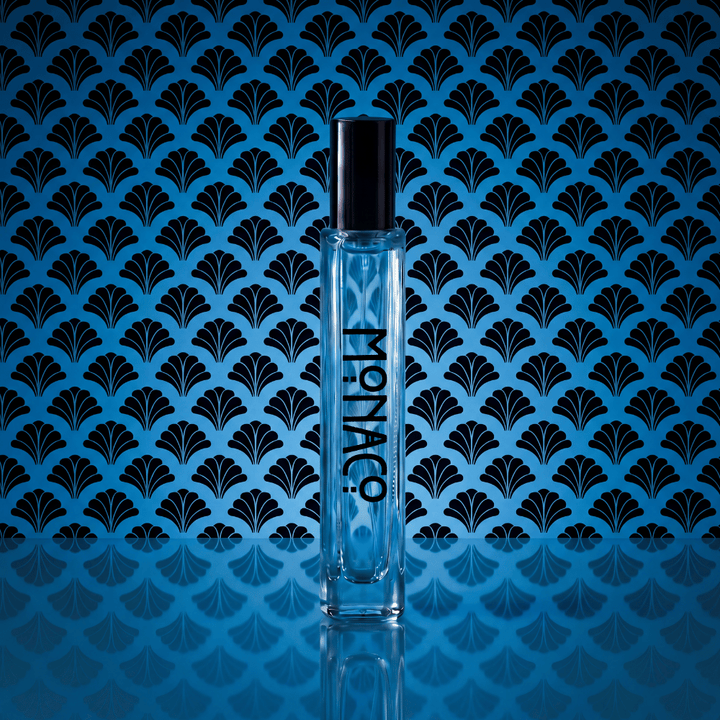 A clear rectangular perfume bottle with a black cap stands centered against a blue background with a repeating black fan-like pattern. The bottle is labeled 'MONACO' vertically.