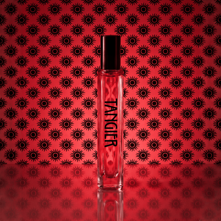A clear rectangular perfume bottle with a black cap stands centered against a red background patterned with repeating black sunburst motifs. The bottle is labeled 'TANKIER' in black text.
