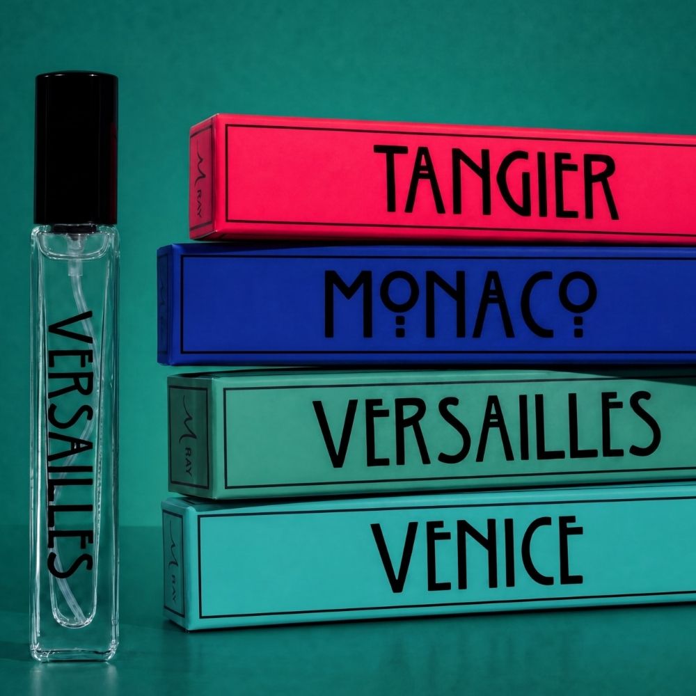 A clear glass perfume rollerball bottle labeled VERSAILLES with a black cap stands beside a stack of four colorful rectangular boxes labeled TANGIER, MONACO, VERSAILLES, and VENICE on a teal background.