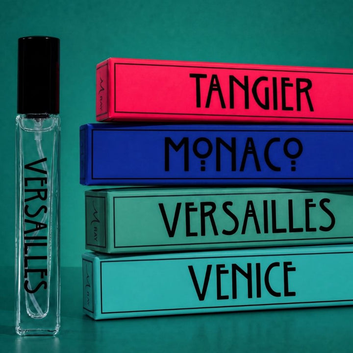 A clear glass perfume rollerball bottle labeled VERSAILLES with a black cap stands beside a stack of four colorful rectangular boxes labeled TANGIER, MONACO, VERSAILLES, and VENICE on a teal background.