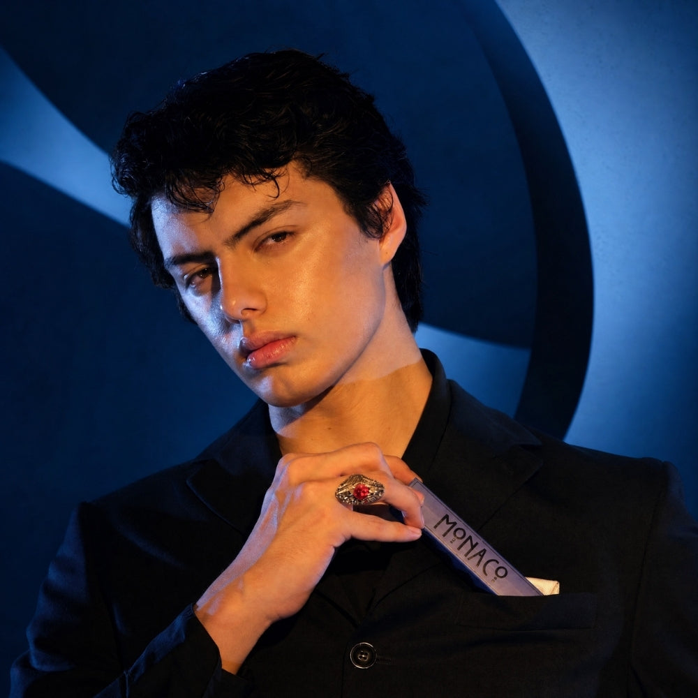 A man in a black suit jacket holding a silver-toned watch with a red gemstone bezel and 'MONACO' strap, posed against a dark blue circular background.