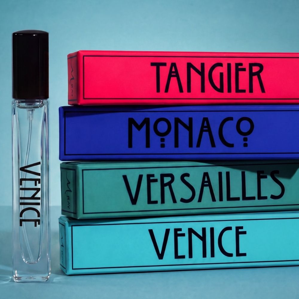 A clear rectangular perfume bottle with a black cap stands beside a stack of four colorful boxes labeled TANGIER, MONACO, VERSAILLES, and VENICE on a light blue background.