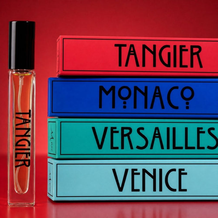 A clear rectangular perfume rollerball bottle with a black cap stands beside a stack of four colorful boxes labeled TANGIER, MONACO, VERSAILLES, and VENICE on a red background.