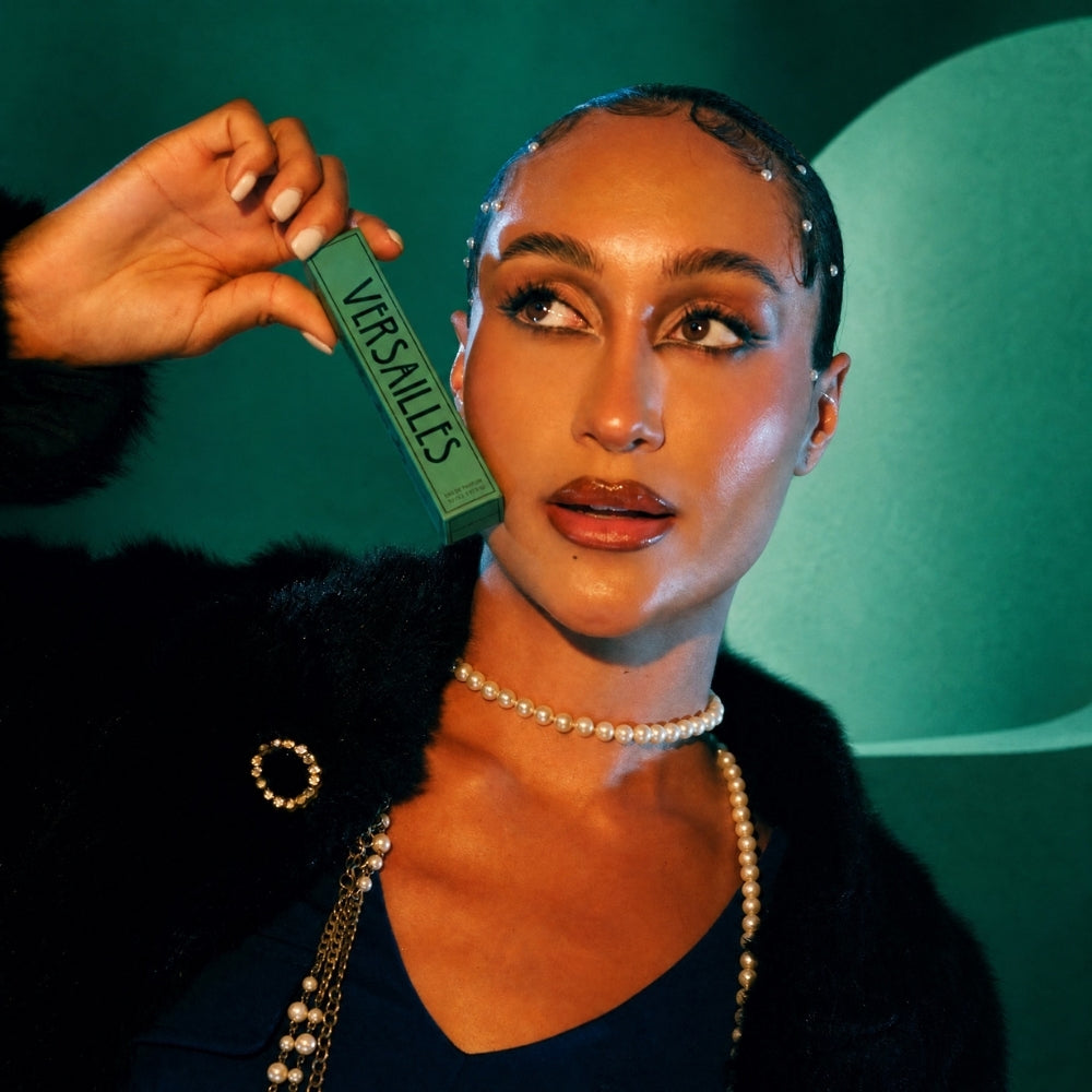 A woman in a black fur-trimmed top and pearl necklace holds up a green Versailles box against a dark green background.
