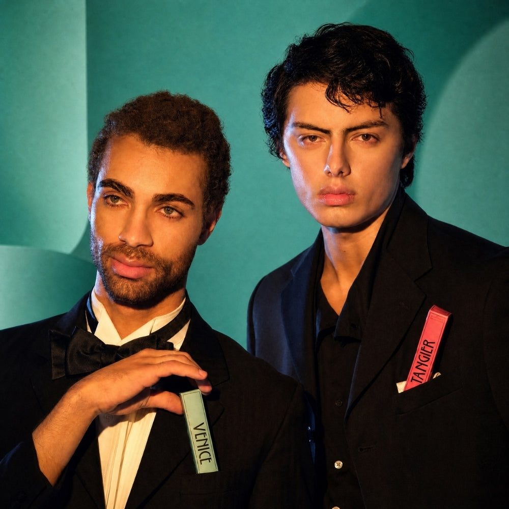 Two men in black formalwear posing against a teal studio backdrop. One wears a black tuxedo jacket with a white shirt and black bow tie, holding a light green tag that reads VENICE.