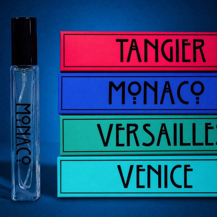 A clear glass perfume rollerball bottle with a black cap stands beside stacked rectangular cards reading TANGIER, MONACO, VERSAILLE, and VENICE on a blue background.
